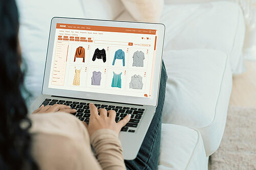 Person sat on a white sofa, back of their shoulder, their hands and their legs visible. The person is using a laptop. On the laptop screen is an online clothes shopping website.